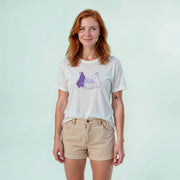 A woman with red hair wearing a white t-shirt with a graphic design and khaki shorts stands against a light blue background.