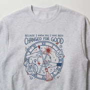 A gray sweatshirt with a graphic design featuring a clock, stars, and various other elements. The text above the design reads 