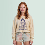A young woman with red hair wearing a beige sweater with a graphic design and denim shorts stands against a light blue background.