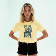 A young woman with blonde hair wearing a yellow t-shirt with a graphic design and black shorts stands with her hands on her hips.