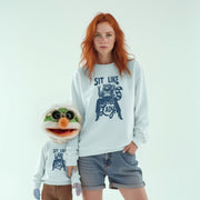 A woman with red hair wearing a white sweatshirt with a graphic design and a smaller version of the same graphic design on the sweatshirt stands next to a smaller, anthropomorphic character wearing a similar design.