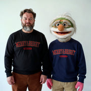 A man in a black sweater with red text and a man in a blue sweater with red text stand next to each other, with a character wearing a green and white mask with red text on it.