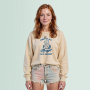 A young woman with red hair wearing a beige sweatshirt with a graphic design and denim shorts stands against a light blue background.