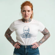 A woman with red hair and tattoos is wearing a white t-shirt with a graphic design and text printed on it.