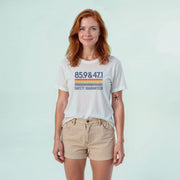 A woman with red hair wearing a white t-shirt with a graphic design and khaki shorts stands against a light blue background.