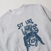 A gray sweatshirt with a graphic design featuring a raccoon sitting in a chair and holding a drink, with the text 
