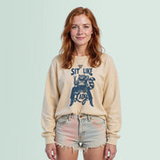 A young woman with red hair wearing a beige sweatshirt with a graphic design and denim shorts stands against a light blue background.