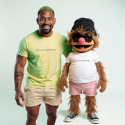 A man in a yellow t-shirt and shorts stands next to a large, furry character wearing sunglasses and a beanie, both with the text 