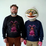 A man in a black sweater with a Christmas-themed design and a man in a blue sweater with a Christmas-themed design stand together, with a character wearing a green and white face mask with a red nose and a pink and purple design on the front.