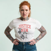 A woman with red hair wearing a white t-shirt with a graphic design and tattoos on her arms stands with her hands on her hips.