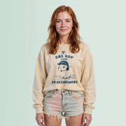 A woman with long red hair wearing a beige sweater with a graphic design and text, standing against a light blue background.