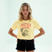 A young woman with blonde hair wearing a yellow t-shirt with a graphic design and black shorts stands with her hands on her hips.