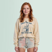 A young woman with red hair wearing a beige sweatshirt with a graphic design and text, standing against a light blue background.