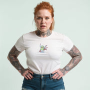 A woman with red hair and tattoos is standing with her hands on her hips, wearing a white t-shirt with a graphic design.