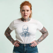 A woman with red hair and tattoos is wearing a white t-shirt with a graphic design and text printed on it.