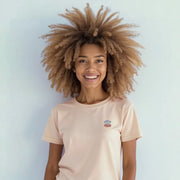 A smiling woman with curly hair wearing a pink t-shirt stands against a white background.