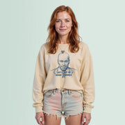 A young woman with red hair wearing a beige sweater with a graphic of a man's face and the text 