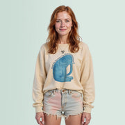 A young woman with red hair wearing a beige sweater with a blue elephant graphic and denim shorts, smiling at the camera.