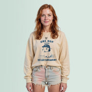 A woman with long red hair wearing a beige sweater with a graphic design and text, standing against a light blue background.