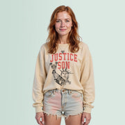 A young woman with red hair wearing a beige sweatshirt with a graphic design and denim shorts stands against a light blue background.