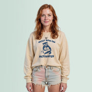 A young woman with red hair wearing a beige sweatshirt with a graphic design and text, standing against a light blue background.