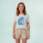 A woman with red hair wearing a white t-shirt with a graphic of a blue elephant and khaki shorts stands against a light blue background.