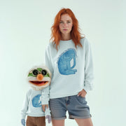 A woman with red hair wearing a white sweatshirt with a blue elephant graphic and jeans stands next to a smaller, green and white stuffed animal.