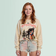 A woman with long red hair wearing a beige sweater with a Christmas-themed design and denim shorts stands against a light blue background.