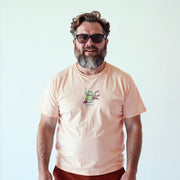 A man with a beard wearing a peach-colored t-shirt with a frog graphic and sunglasses is smiling at the camera.