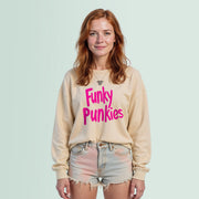 A young woman with red hair wearing a beige sweater with pink text and denim shorts stands against a light green background.