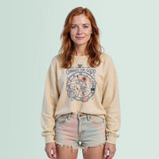 A young woman with red hair wearing a beige sweatshirt with a graphic design and denim shorts stands against a light blue background.