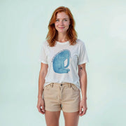 A woman with red hair wearing a white t-shirt with a graphic of a blue elephant and khaki shorts stands against a light blue background.