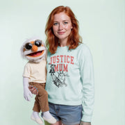 A woman with red hair is holding a large, white, cartoonish stuffed animal with sunglasses on its head. The stuffed animal is wearing a shirt with the text 