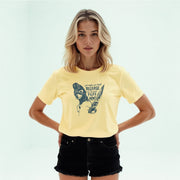 A young woman with blonde hair wearing a yellow t-shirt with a graphic design and black shorts stands with her hands on her hips.