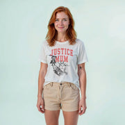 A woman with red hair wearing a white t-shirt with a graphic design and khaki shorts stands against a light blue background.