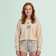 A young woman with red hair is standing against a light blue background, wearing a beige sweater with a frog graphic and denim shorts.